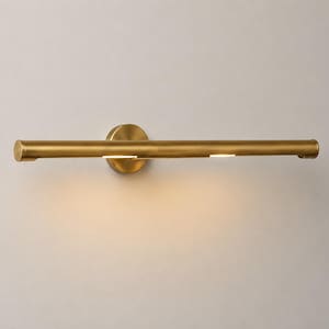 May include: A horizontal wall-mounted light fixture with a cylindrical brass-toned metal body. The design features a circular mounting plate at the center and a long, slender tube that emits a soft, warm glow from two small openings along the bottom.