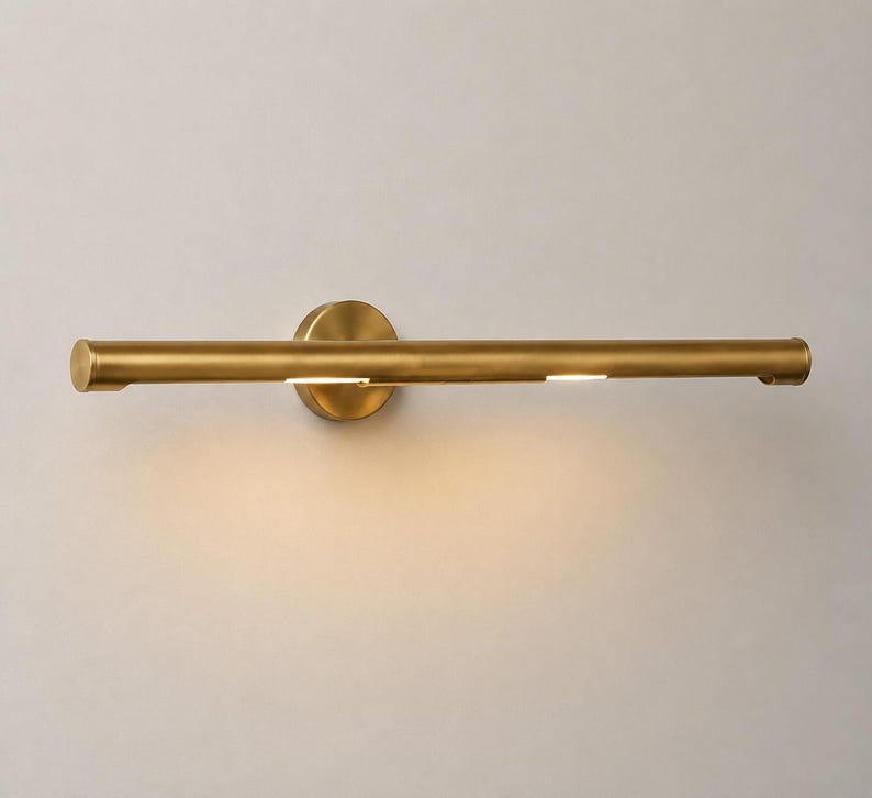May include: A horizontal wall-mounted light fixture with a cylindrical brass-toned metal body. The design features a circular mounting plate at the center and a long, slender tube that emits a soft, warm glow from two small openings along the bottom.