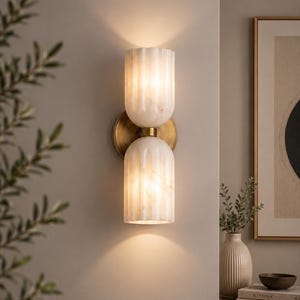 May include: A wall-mounted light fitting featuring two vertically stacked, fluted, off-white glass shades with a marbled texture. The shades are connected by a central brass-toned metal component, which is attached to a circular brass-toned wall plate.