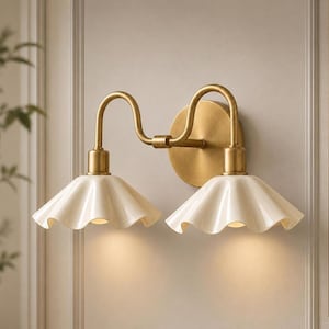 May include: A double wall sconce featuring a brushed brass finish mounting plate and two curved arms. The fixture includes two cream-colored, fluted, bell-shaped shades that direct light downward, creating a soft glow from the light source.