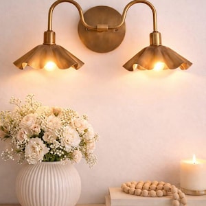May include: A brass wall sconce with two lights, each with a ruffled shade. The sconce is mounted on a wall above a shelf with a white vase of flowers, books, a candle, and decorative beads.