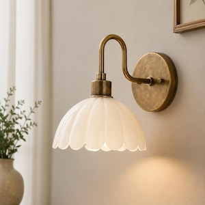 Peut inclure: Une applique murale composée d'un bras en métal courbé au fini laiton antique et d'une plaque murale circulaire. La lampe comprend un abat-jour en verre blanc festonné en forme de pétale de fleur, diffusant la lumière vers le bas.