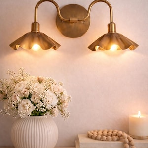 May include: A brass wall sconce with two lights, each with a ruffled shade, illuminates a soft, warm glow. A white vase with a bouquet of flowers sits on a wooden shelf, alongside books and a candle.