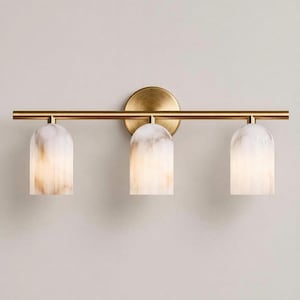 May include: A three-light vanity fixture with a brass finish. The fixture features three white and tan marble-look shades. The shades are dome-shaped and hang from a horizontal brass bar. The fixture is mounted on a round brass backplate.