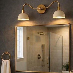 May include: A bathroom vanity set featuring a gold-framed mirror, a double-light fixture, and a gold tap. The vanity includes a marble countertop, a towel ring, and a tray with toiletries. The overall aesthetic is modern and elegant.
