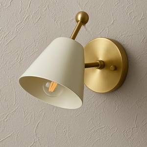 Può includere: Applique da parete color crema con paralume conico e braccio e base color ottone. L'applique presenta una lampadina visibile ed è progettata per il montaggio a parete. L'apparecchio ha un'estetica moderna e minimalista.