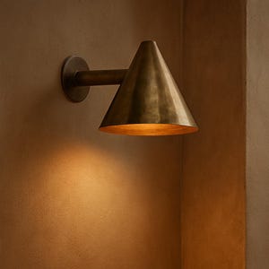 May include: A wall-mounted sconce featuring a conical metal shade with a brushed brass finish. The fixture is attached to the wall by a circular base and a horizontal arm, casting a warm, downward-facing light onto the wall surface below.