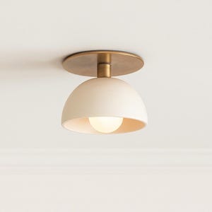 May include: A contemporary ceiling light. It features a cream-coloured, dome-shaped shade and a brass-coloured base. The light bulb is visible inside the shade. The fixture is mounted on a round, brass-coloured plate.