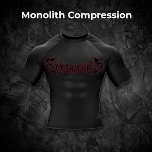 Op de afbeelding: Zwart compressieshirt met korte mouwen en een ronde hals. Het shirt heeft de rode tekst "GYMSHACKLES" over de borst. De woorden "Monolith Compression" staan bovenaan de afbeelding.