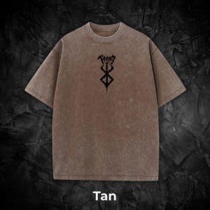 May include: Tan-colored t-shirt with a crew neck and short sleeves. The shirt features a black graphic design on the front, along with Japanese text. The word "Tan" is printed at the bottom of the image.