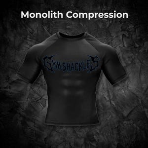 Op de afbeelding: Zwart compressieshirt met korte mouwen en de tekst "GYMSHACKLES" in een blauw, gestileerd lettertype. Het shirt wordt getoond tegen een donkere, getextureerde achtergrond, met de woorden "Monolith Compression" bovenaan.
