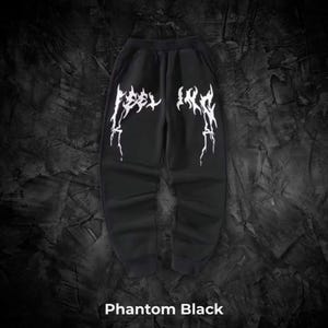 Peut inclure: Pantalon de survêtement noir avec l'inscription blanche "FEEL IRK" dans un style gothique. Le pantalon est de couleur noir fantôme et a une coupe décontractée. Le fond est sombre et texturé.