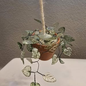 May include: A hanging terracotta pot with clear glass leaves and stems. The leaves are a light green color and are attached to the pot with dark wire. The pot is suspended by a natural fiber rope.