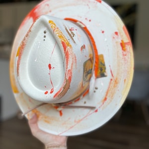May include: A white cowboy hat with orange and red paint splatters. The hat has a brown leather band and a small, colorful patch on the side.