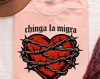 Może przedstawiać: Jasnoróżowy t-shirt z grafiką czerwonego serca owiniętego czarnym drutem kolczastym. Nad sercem znajduje się napis "chinga la migra" wykonany czarną, stylizowaną gotycką czcionką. Wzór ma postarzany, przetarty wygląd.