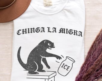 Może przedstawiać: Biały t-shirt z krótkim rękawem z czarnym nadrukiem graficznym przedstawiającym stworzenie stojące na małej platformie i sięgające po słoik z napisem ICE. Nad grafiką znajduje się tekst CHINGA LA MIGRA wydrukowany pogrubioną, czarną czcionką gotycką.