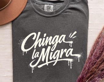 Może przedstawiać: Ciemnoszary t-shirt z krótkim rękawem z białym napisem w stylu graffiti Chinga la Migra. Koszulka ma okrągły dekolt i metkę marki Comfort Colors po wewnętrznej stronie kołnierza. Wzór posiada efekt ściekającej farby w sprayu.