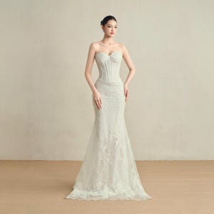 May include: A white lace wedding dress with a strapless design and a fitted bodice. The dress has a mermaid style skirt and is adorned with delicate floral lace. The model is standing in front of a plain background.