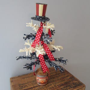 May include: A patriotic decorative tree with a red, white, and blue color scheme. The tree is adorned with red star-patterned ribbon, white and blue accents, and a miniature Uncle Sam hat. It sits atop a wooden crate.