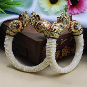 Peut inclure: Une paire de bracelets dorés avec des extrémités en forme de paon ornées d'émail blanc, rouge et vert et de pierres transparentes. Les anneaux circulaires sont recouverts de rangées de petites perles blanches, créant un aspect texturé uniforme.
