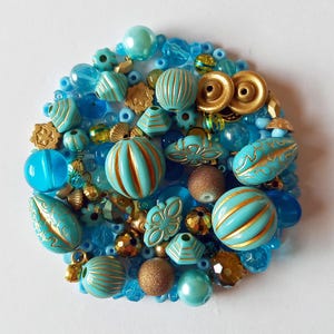 May include: A circular arrangement of various beads in shades of turquoise, blue, and gold. The beads include round, striped, and textured shapes, creating a decorative display. Some beads have gold accents.
