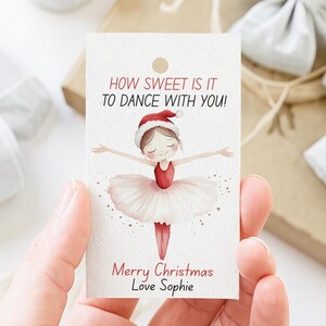 May include: A rectangular white gift tag with a hole at the top. It features a watercolor illustration of a ballerina wearing a red leotard, white tutu, and Santa hat. The text reads: HOW SWEET IS IT TO DANCE WITH YOU! Merry Christmas Love Sophie.
