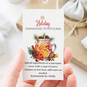 May include: A rectangular white tag featuring the text Holiday SIMMERING POTPOURRI, a watercolor illustration of a glass jar filled with orange slices, cranberries, and cinnamon sticks, and instructions to add 2 cups of water and simmer.