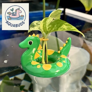 Könnte beinhalten: Ein grüner und gelber Drachen-Pflanzenschwimmer mit einem Pflanzenstiel in der Mitte. Der Schwimmer hat gelbe Punkte und Stacheln. Das Logo "AQUABUDS" befindet sich in der oberen linken Ecke und zeigt ein Einhorn in einem Schwimmer.