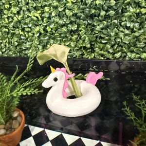 Può includere: Un vaso a forma di unicorno in ceramica bianca con criniera e coda rosa e un corno giallo. Il vaso contiene uno stelo di pianta verde con una grande foglia. Lo sfondo presenta una parete verde di fogliame.