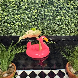 Op de afbeelding: Een felroze opblaasbare flamingo zwemband met gele snavel en oogdetails. Een groen en wit blad is aan de band bevestigd. De achtergrond heeft een groene muur van gebladerte en een zwart-wit geblokt patroon.