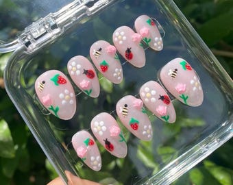 May include: A set of ten artificial nails with a light pink base. Each nail is decorated with a combination of white flowers, pink flowers, red ladybugs, yellow bees, and red strawberries. The nails are in a clear plastic case.
