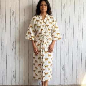 May include: A white robe featuring a repeating pattern of mustard yellow tigers. The robe has three-quarter length sleeves, a V-neckline, a matching fabric tie belt at the waist, and two front patch pockets. The garment falls to mid-calf length.