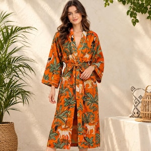 May include: A long-sleeved orange robe featuring a vibrant jungle-themed print with green palm leaves, white leopards, and black toucans. The garment includes a matching fabric tie belt at the waist, a V-neckline, and white piping along the front edges.