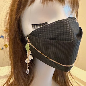 Op de afbeelding: Zwart gezichtsmasker met een gouden ketting en bloemenbedel. De bedel heeft witte bloemen, een groen blad en een heldere kristallen hanger. Het masker wordt gedragen op een mannequinhoofd met bruin haar.