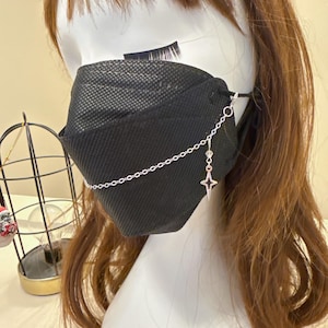 May include: Black face mask with a silver chain and star charm. The mask is made of a textured material and is worn on a mannequin head with brown hair. The chain is attached to the mask with small hoops.