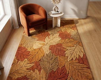 May include: A rectangular rug featuring a leaf design in autumnal shades of orange, brown, and beige. The rug is placed on a wooden floor, with a rust-coloured armchair and a small side table with a vase and dried flowers in the background.