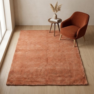A high-angle room view of a soft, solid terracotta plush rug styled in a minimalist space next to a matching rust-colored armchair and a small wooden side table with a vase.