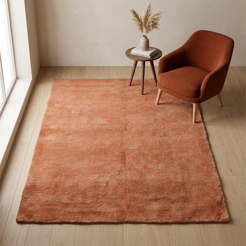 A high-angle room view of a soft, solid terracotta plush rug styled in a minimalist space next to a matching rust-colored armchair and a small wooden side table with a vase.