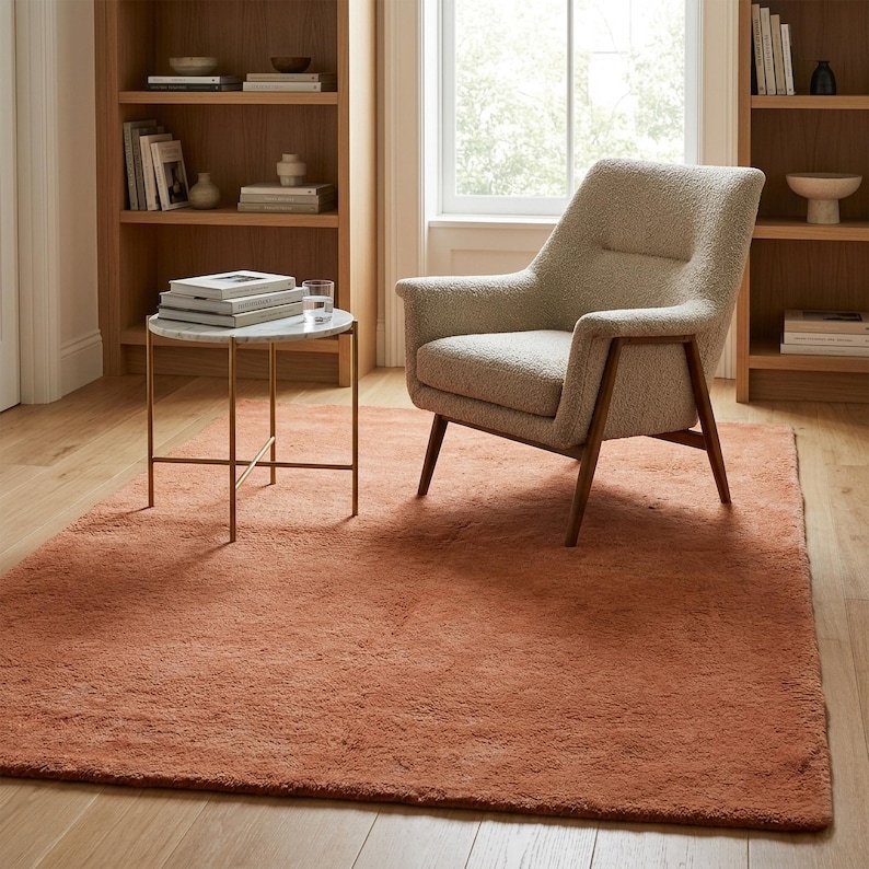 A lifestyle view of a thick, terracotta-orange accent rug placed in a cozy reading nook on hardwood flooring, styled with a modern bouclé armchair, small marble table, and built-in wooden bookshelves.