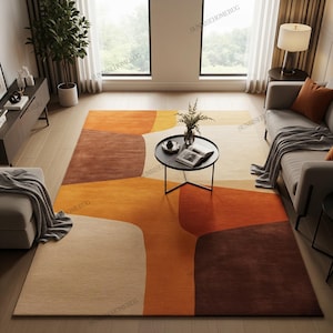 May include: A large, abstract rug with a geometric design in shades of orange, brown, and beige. The rug features a modern, contemporary style with soft, curved shapes. The rug is placed in a living room setting with a sofa, coffee table, and natural light.