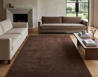 May include: A large, rectangular, solid dark brown area rug with a plush, textured pile. The rug is positioned centrally on a light-colored wooden floor, extending from the foreground toward a brown sofa and a wooden coffee table in the room.