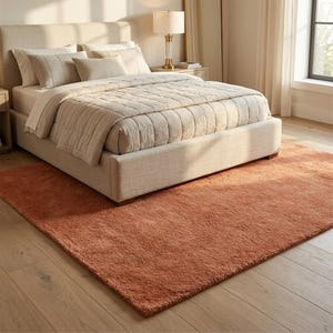 A perspective shot of a thick, solid terracotta area rug positioned under a modern beige upholstered bed frame with neutral linens, casting warm sunlight across light wood flooring in a bright bedroom setting.