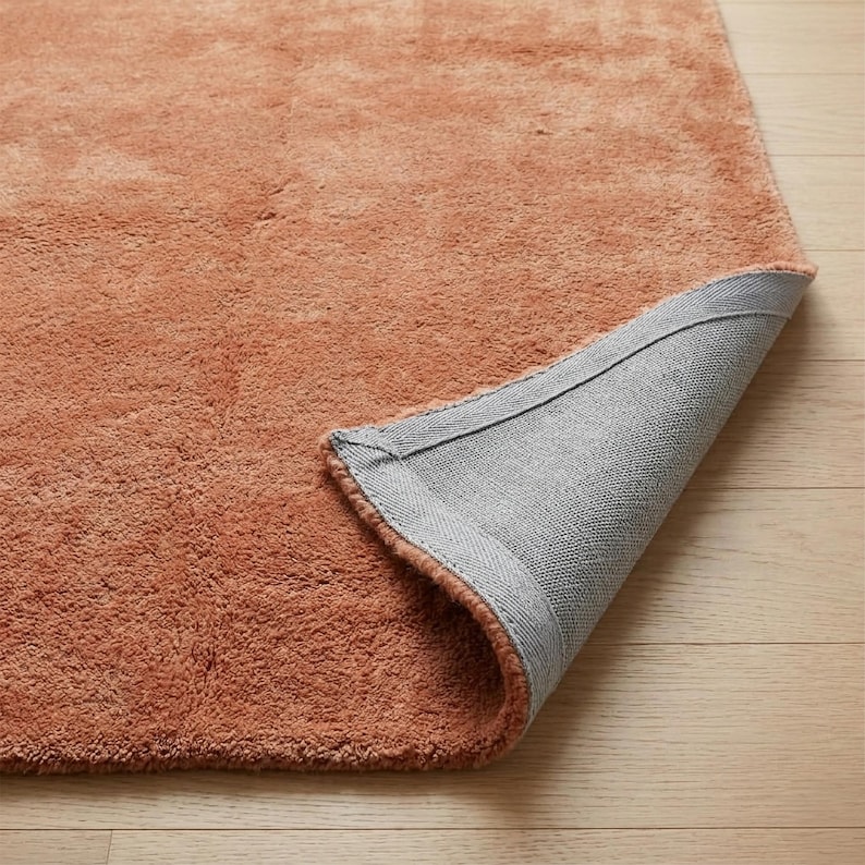 A close-up detailed shot of a plush, terracotta-colored area rug showing a corner folded over to display its durable, non-slip gray woven backing on a light wood floor.