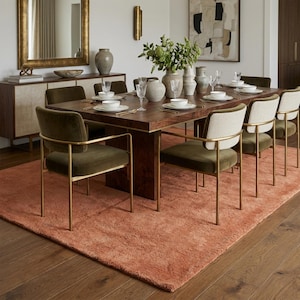 A wide-angle lifestyle view of a large terracotta-colored plush rug layered underneath a long, dark wood dining table set for eight, surrounded by elegant olive green velvet dining chairs in a modern dining room.