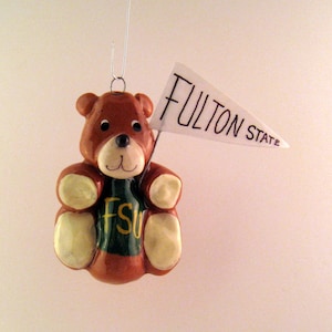 May include: A brown teddy bear ornament with a green shirt that says "FSU" on it. The bear is holding a white pennant that says "Fulton State".