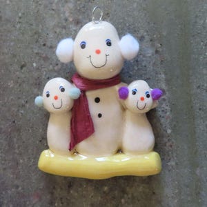 May include: A ceramic ornament featuring a large snowman with white pom-pom earmuffs and a maroon scarf, flanked by two smaller snowmen. The smaller snowmen have light blue and purple pom-pom earmuffs. All figures stand on a yellow base.