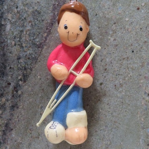 May include: A ceramic ornament depicting a person with brown hair, wearing a red shirt and blue pants. The figure is holding a pair of cream-colored crutches and has a bandaged foot. The ornament is designed to hang.