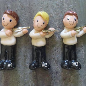May include: Three ceramic ornaments shaped like figures playing violins. Each figure wears a white shirt and black pants. Figure #1 has brown hair, figure #2 has yellow hair, and figure #3 has brown hair. Each has a small metal loop at the top.
