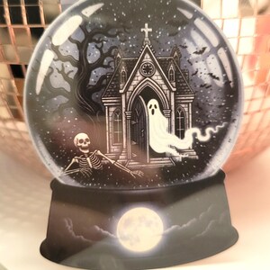 May include: A Halloween-themed snow globe featuring a spooky scene. Inside the glass sphere, a white ghost emerges from a haunted chapel, with a skeleton and a full moon at the base. The background includes a bare tree and bats.