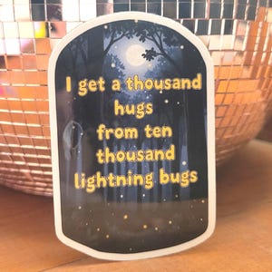 Op de afbeelding: Een sticker met een donkere bosscène, een volle maan en de tekst "I get a thousand hugs from ten thousand lightning bugs" in het geel. De sticker heeft een witte rand en een afgeronde bovenkant.